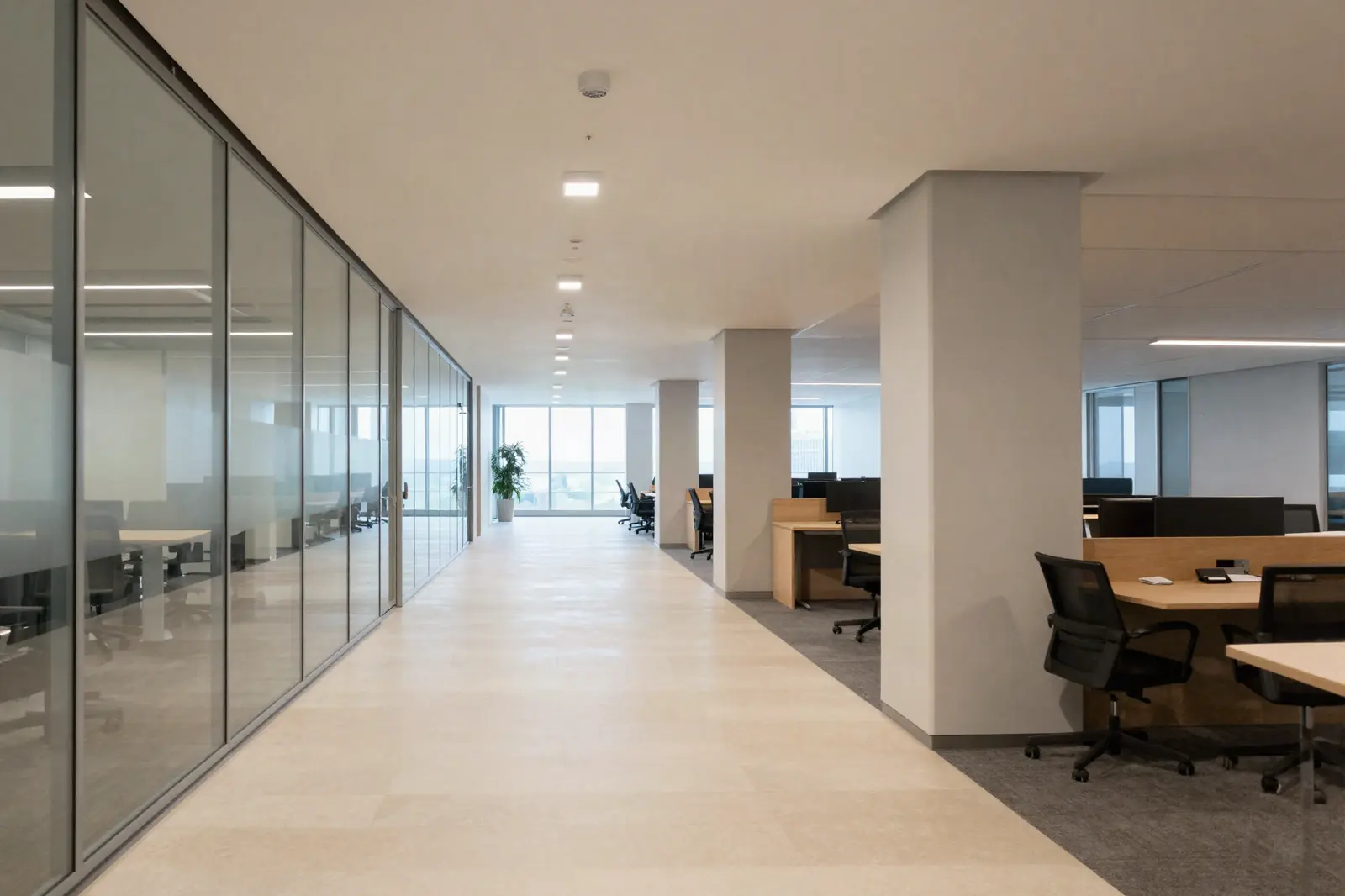 A wide primary circulation route down an office floor, glass-fronted meeting rooms along one side and bench desking beyond on the other, daylight at the far end.